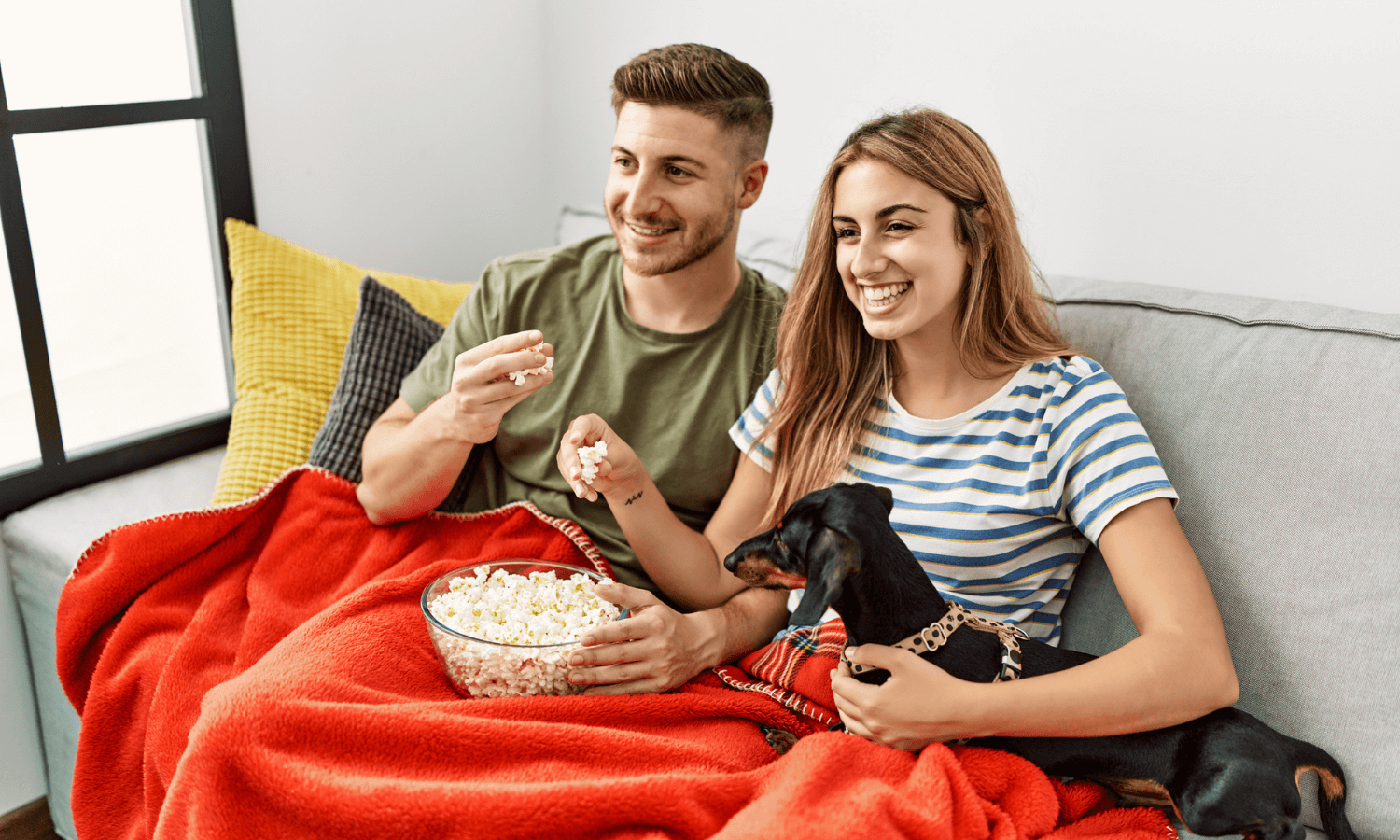 Ein Paar mit Hund sitzt auf dem Sofa und schaut einen Film. Sie kuscheln sich in eine Decke und essen zusammen Popcorn. © iStock.com