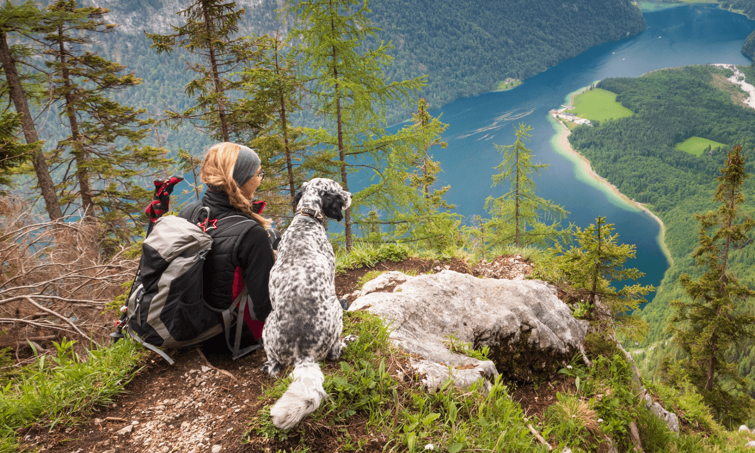 Eine Frau geht während der Osterfeiertage mit ihrem Hund in den Bergen wandern. Sie sitzen nebeneinander auf einem Felsvorsprung und blicken hinunter in ein Tal mit einem See. © iStock.com