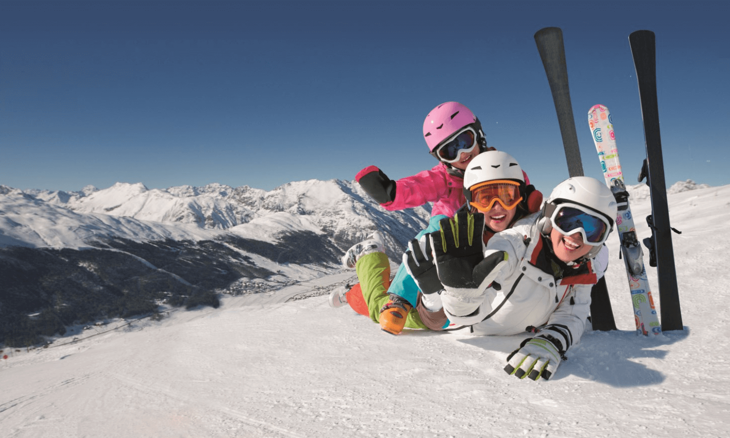 Skifahren können Kinder in fast allen Skigebieten lernen. Auf dem Bild sieht man drei Kinder mit Skiausrüstung, die auf dem Bauch im Schnee liegen und für das Foto posieren. © iStock