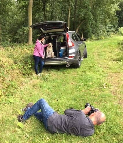 photographer lying in the grasss taking photo of woman standing next to SUV with dog in the trunk